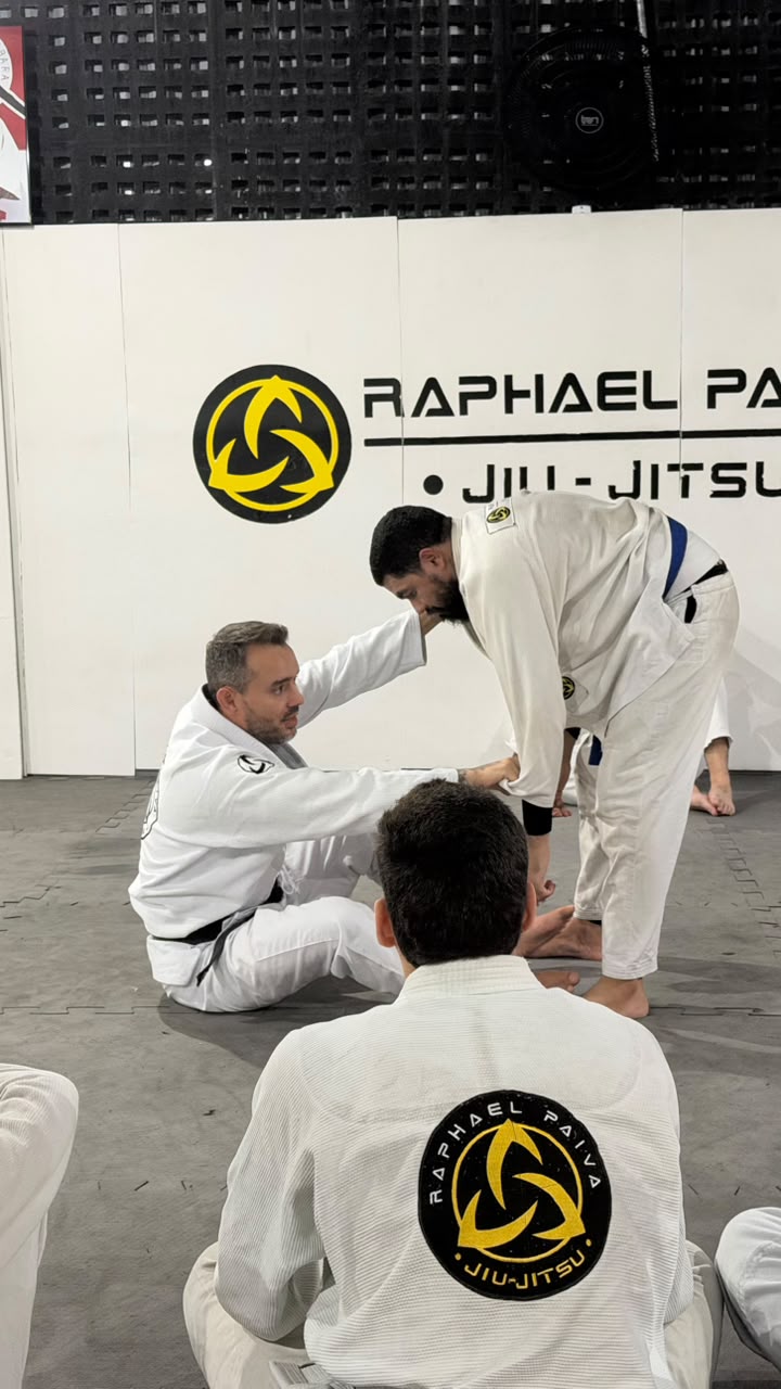 Professor demonstrando a técnica diante da parede com o logo do Raphael Paiva Jiu-Jitsu